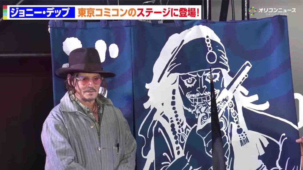 Johnny Depp standing next to a noren curtain with an illustration of Jack Sparrow by Eiichiro Oda.